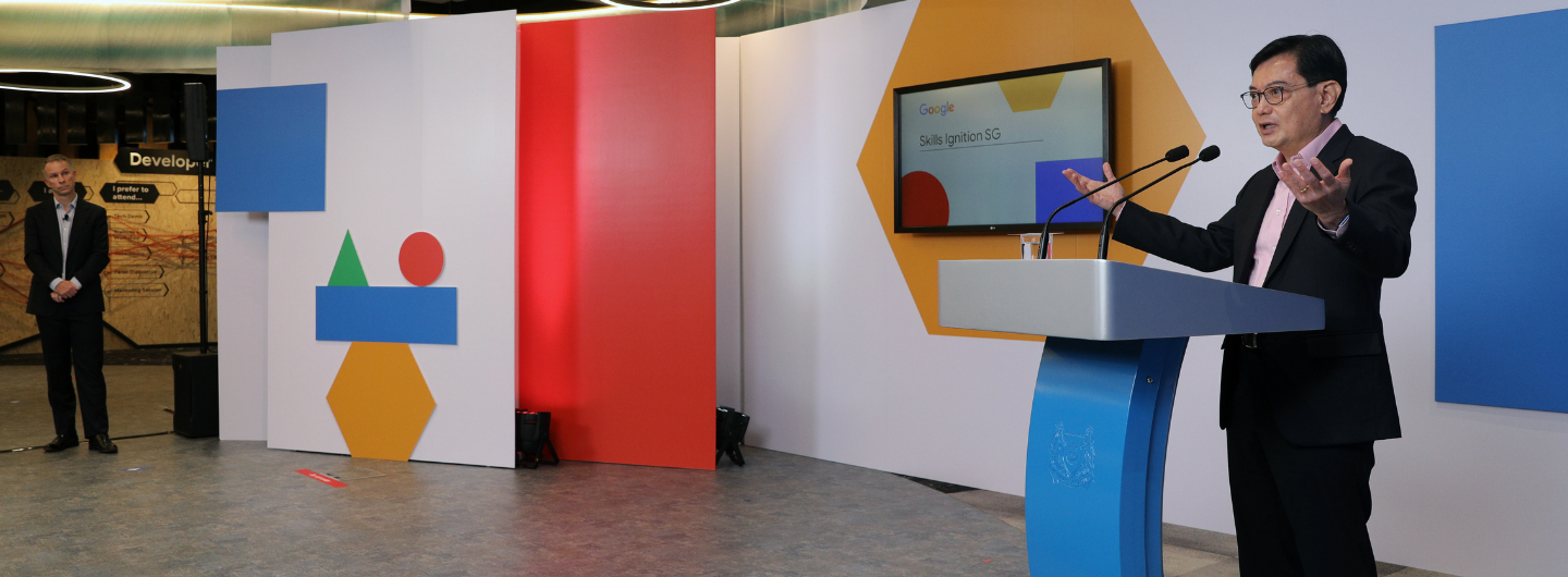 Speaker at podium with Google display, shapes, and wall panels, another person stands on the left.